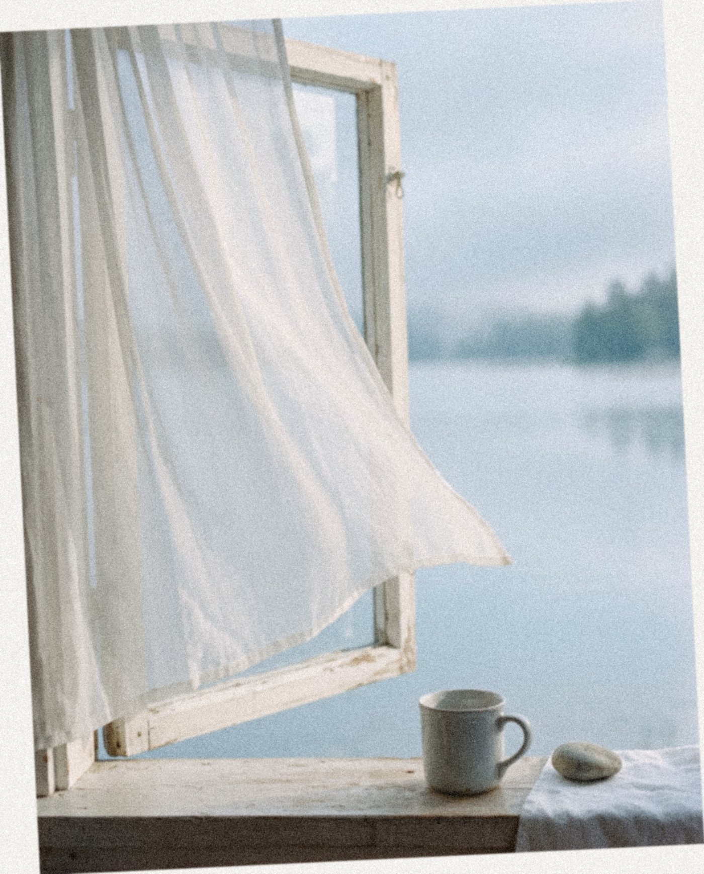A sheer white curtain lifting at an open window with a lake beyond