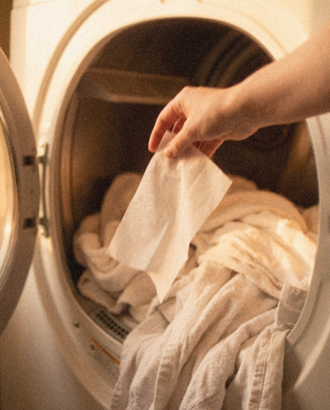 A hand placing a white dryer sheet onto towels in an open dryer
