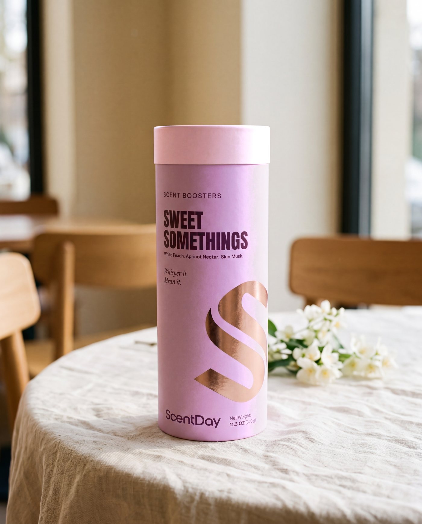 Sweet Somethings scent booster tube in peach and pink on a café table beside pink flowers